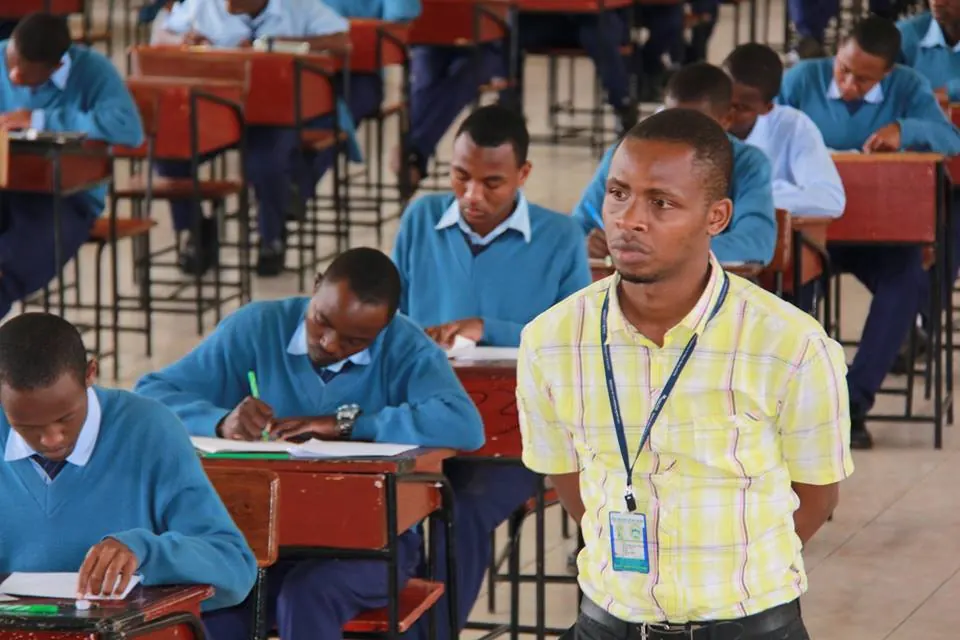 Students in blue school jumpers writing an exam at their desks while an invigilator watches the room