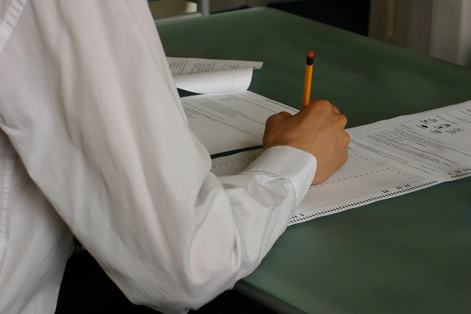 A student in a white shirt writes an exam answer with a pencil at a desk in a classroom