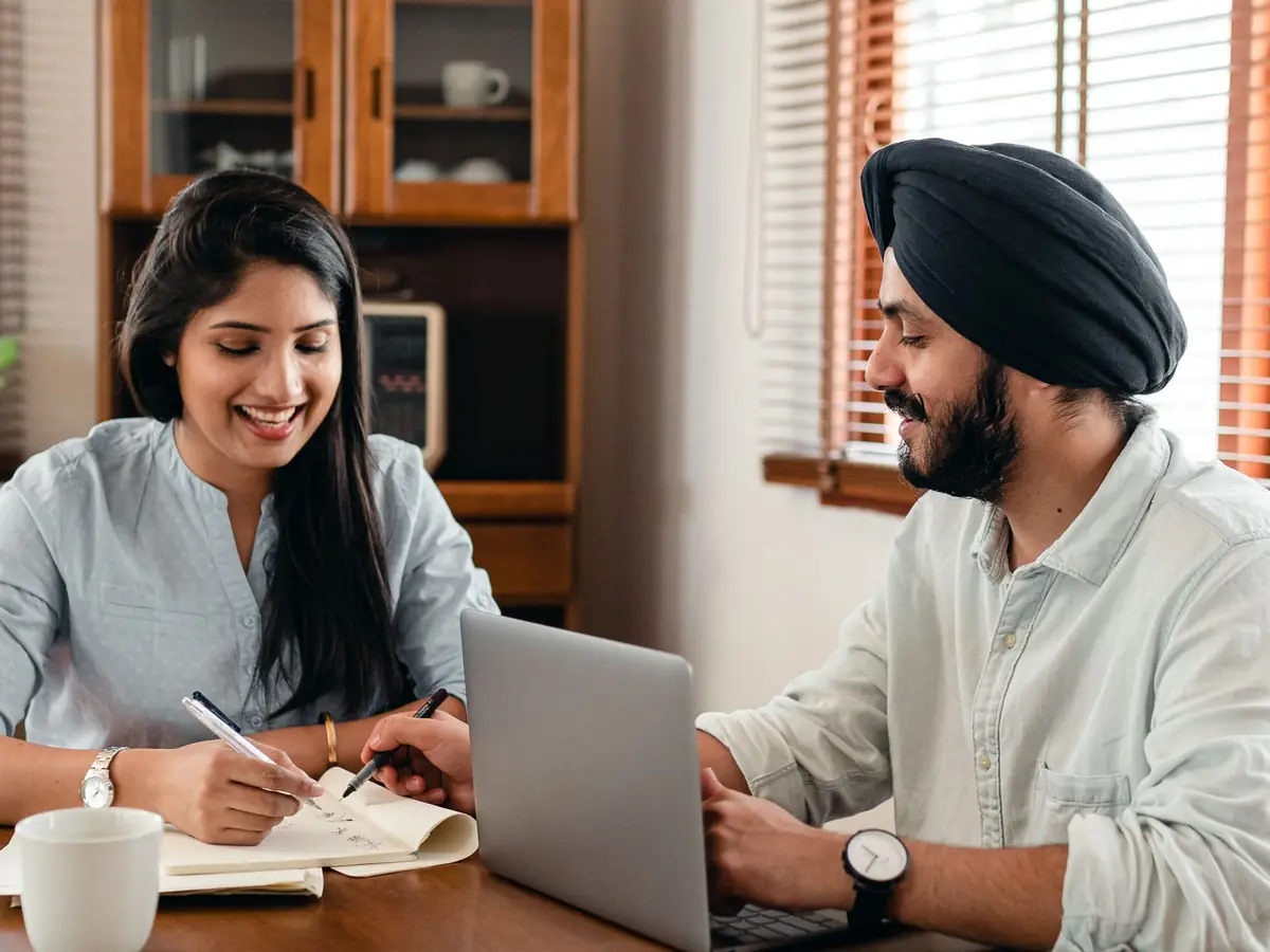 A smiling student takes notes at a table while a tutor works beside her on a laptop