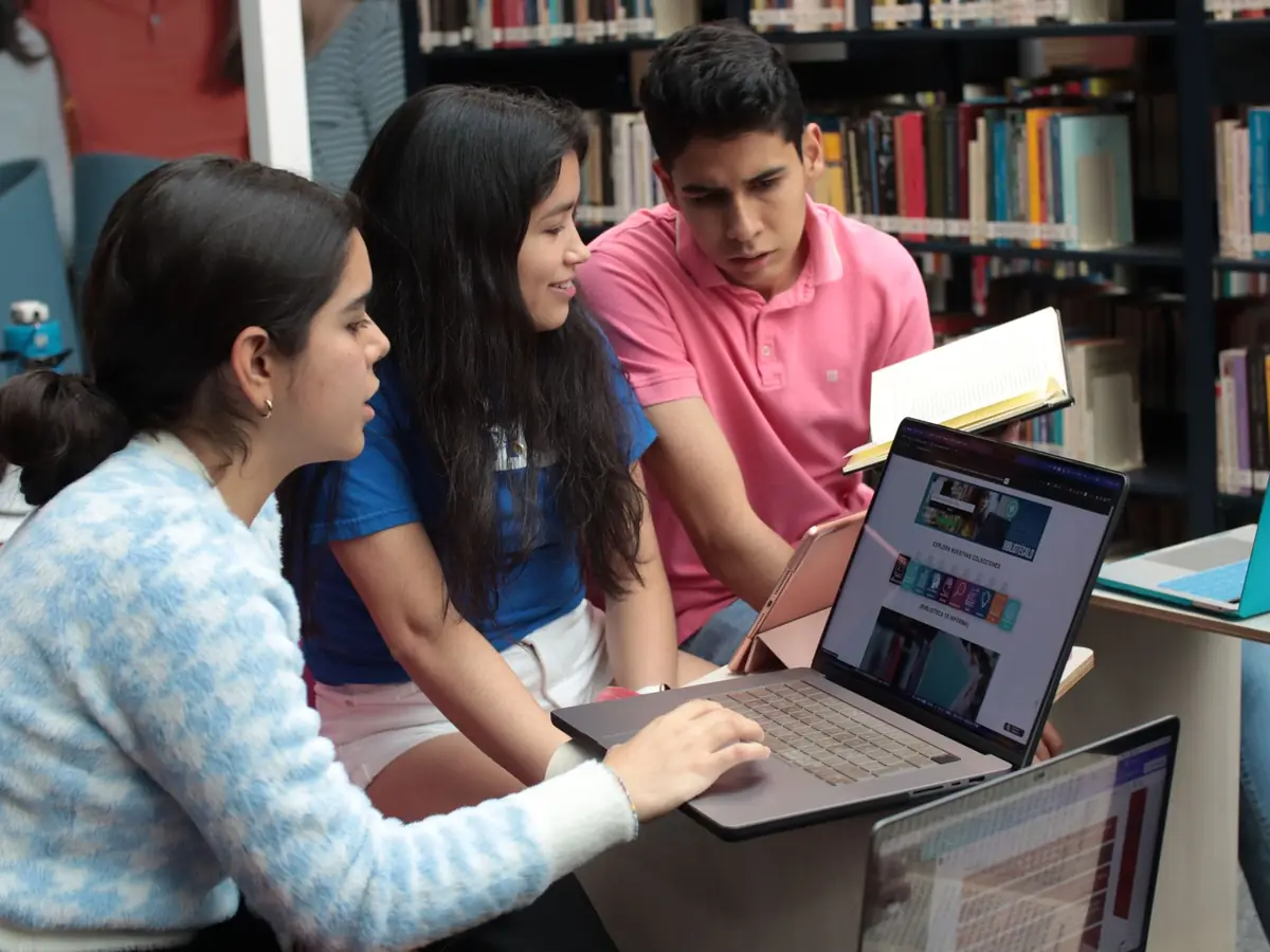 Three young students study together in a library, discussing what is on a laptop