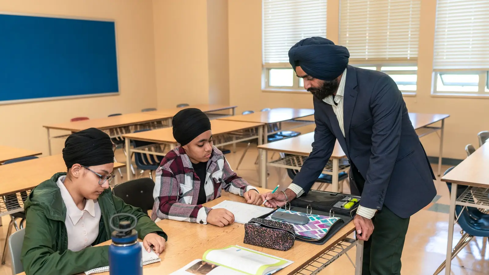 A teacher leans over a desk to help two students with their work in a bright classroom